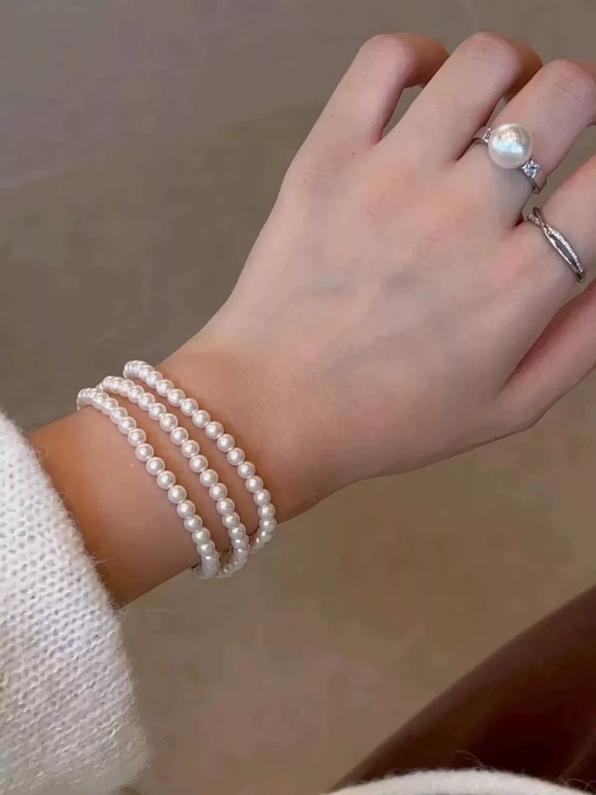 Three-strand synthetic pearl bracelet worn on a wrist, with pearl and silver-colored rings on the hand.