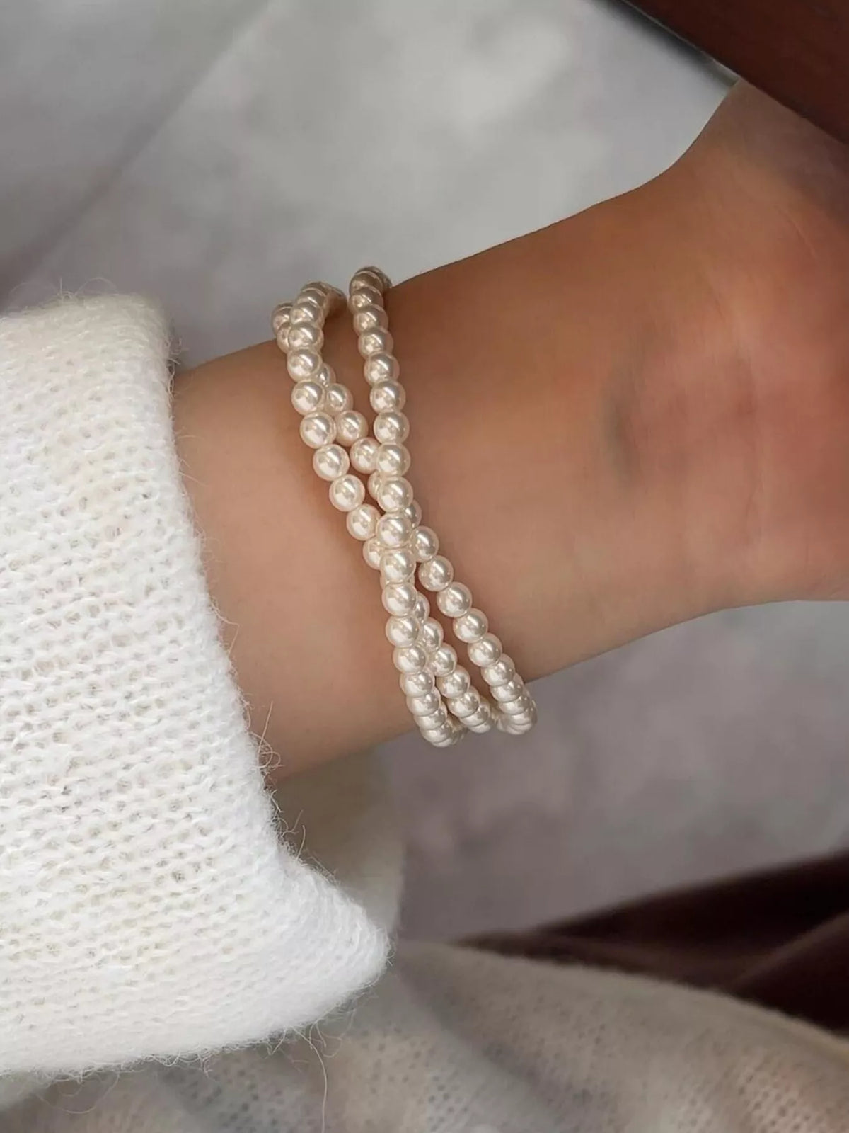 Close-up side view of three synthetic pearl strands around a wrist beside a white knit sleeve.